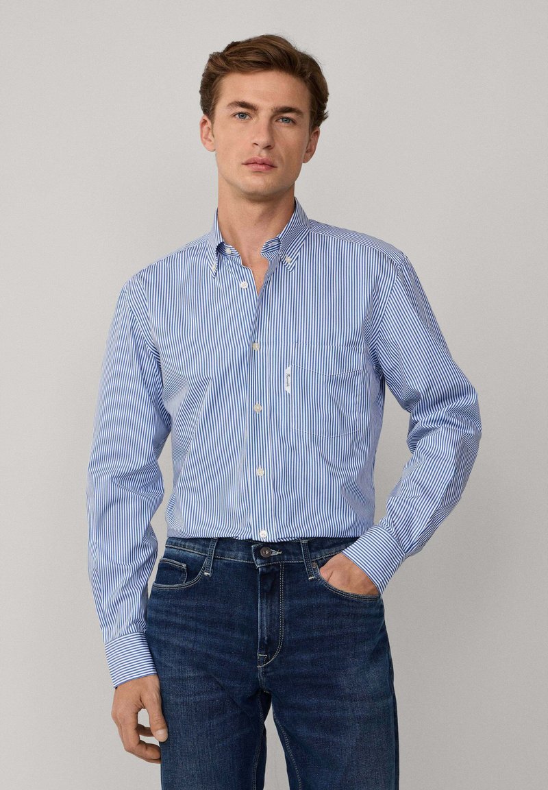 Camisa de botones a rayas azul y blanca con mangas largas y un bolsillo en el pecho, combinada con jeans oscuros. Material de algodón, diseño de cuello clásico.