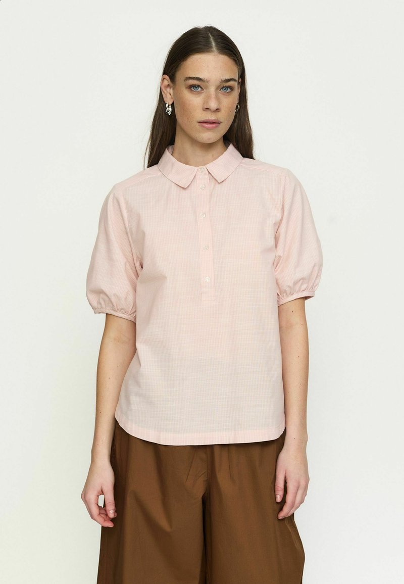 Femme aux longs cheveux bruns portant une chemise rose clair à manches courtes avec col et un pantalon marron pantalon large, debout devant un fond uni.