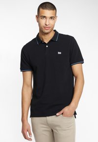 Polo noir avec accents bleus, présentant une patte de boutonnage à trois boutons et un logo brodé sur la poitrine, associé à un pantalon beige.