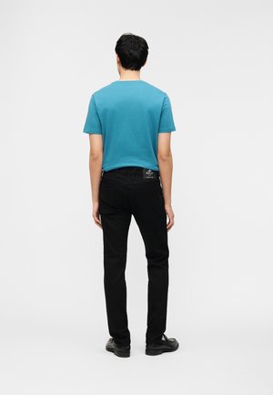 Homme debout de dos, portant une chemise bleue à manches courtes, un pantalon noir avec une étiquette visible, et des chaussures noires sur fond blanc.