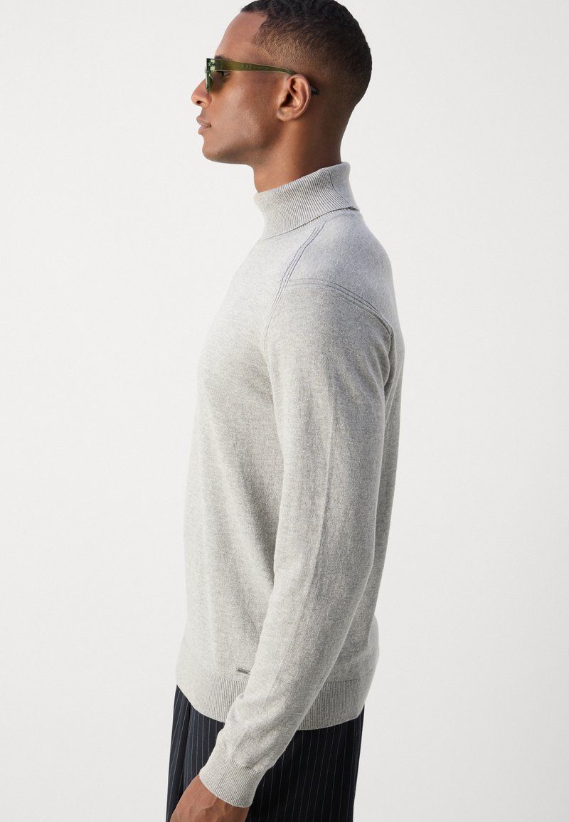 Un chandail à col roulé gris clair en tricot, avec des manches ajustées, des poignets côtelés et des détails de couture subtils. Porté avec un pantalon à rayures verticales foncées.