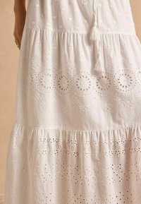 Witte katoenen jurk met geborduurde bloemmotieven, ooglet-details en gelaagde tiers met een gerufszoom. Twee kwastjes bij de halslijn.