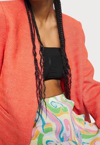 Blazer corail à motif chevrons avec épaules structurées, associé à un top court noir et une jupe multicolore à motif tourbillonnant.