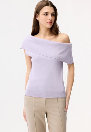 Vrouw in een lichtpaarse off-shoulder top en beige broek, staand met één hand in de zak, kijkt iets naar rechts.