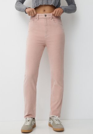 Person iført lyserøde bukser med høj talje og grå sneakers, hænder justerer linningen, grå cropped sweater, ens hvid baggrund.