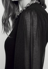 Svart topp med ribbad kropp, genomskinliga långärmar och en metallkedja som accent vid halsringningen, vilket framhäver en strukturerad design.