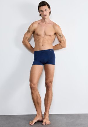 Mężczyzna bez koszulki z tatuażami stoi boso oparty o białą ścianę, mając na sobie granatowe bokserki.