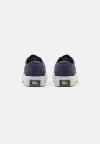 Tumedast sinised Lacoste kanvasest spordijalanõud vaadet tagant, näidates valgeid kummist taldi ja rohelisi Lacoste logo silte.