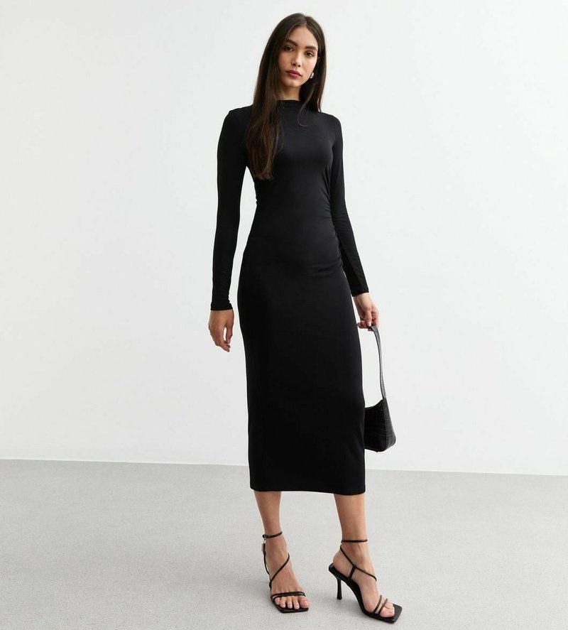 Robe noire moulante à manches longues en tissu extensible, longueur genou avec un décolleté rond. Le modèle tient un petit sac à main noir et porte des talons avec des lanières.