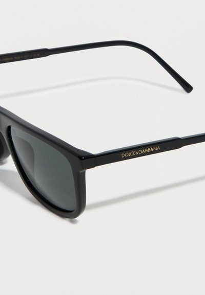 Μαύρα ορθογώνια γυαλιά ηλίου με σκούρους φακούς και το λογότυπο Dolce & Gabbana σε χρυσό στο μπράτσο του βραχίονα, σε λευκό φόντο.