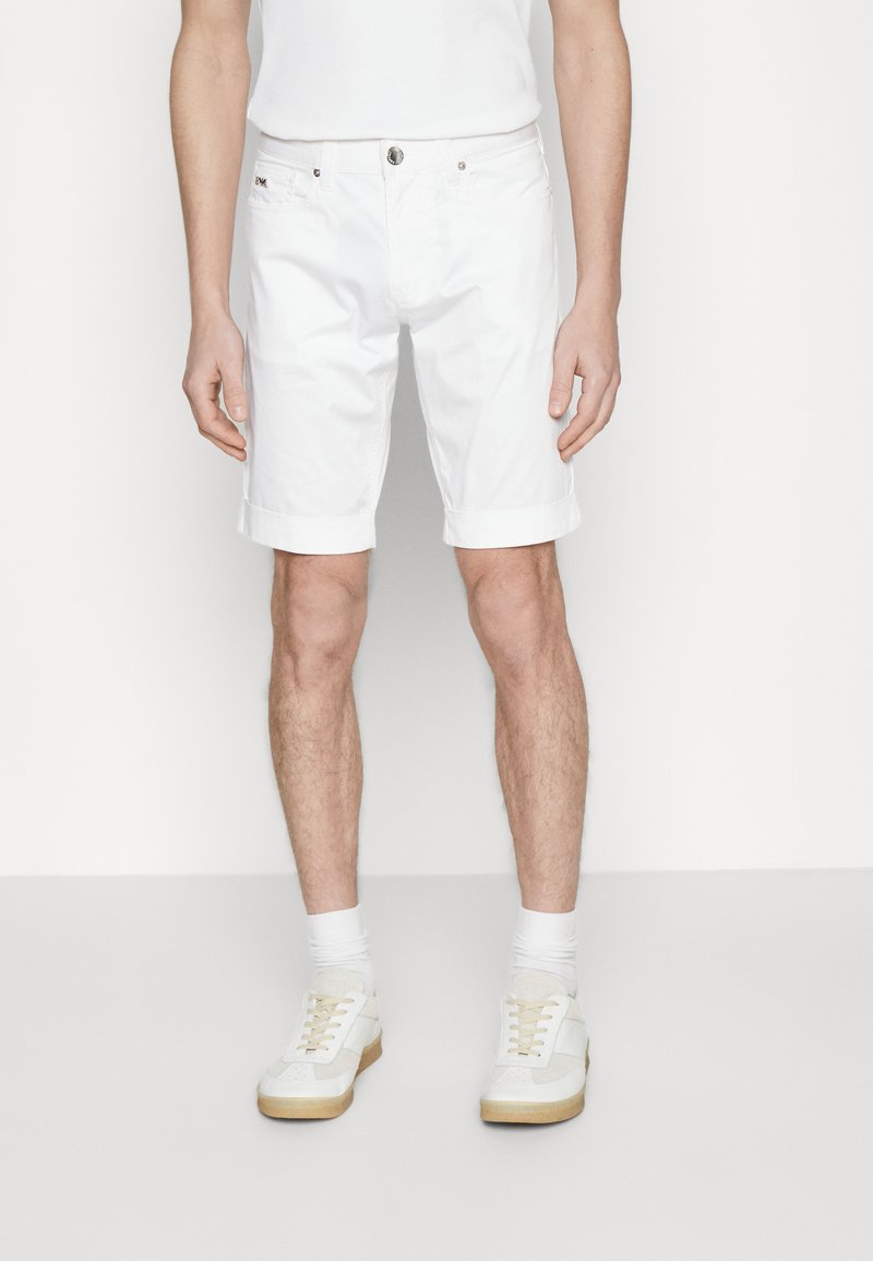 Hvite bomullsshorts med rullet kant, med knappelukking foran og lommer, satt sammen med hvite joggesko og sokker.