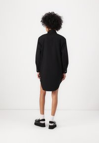 Abito camicia oversize nero con maniche lunghe, polsini abbottonati, spacchi laterali e colletto. Abbinato a scarpe flatform nere e calzini bianchi.