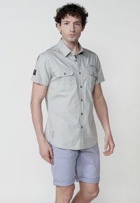 Camicia abbottonata a maniche corte grigio chiaro con due tasche al petto, bottone logo nero sulla manica e un design con orlo curvo.