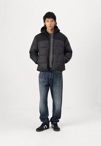Černá péřová bunda s horizontálním prošíváním, spárovaná se šedým tričkem a roztrhanými modrými džíny. Outfit doplňují černé tenisky.