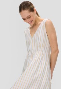 Robe blanche avec des rayures dorées verticales, décolleté en V, sans manches, taille cintrée et jupe fluide. Texture de tissu lisse.