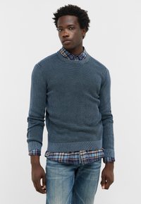 Niebieski sweter o fakturze z okrągłym dekoltem, noszony na koszuli w kratę z guzikami. Posiada długie rękawy i dopasowaną talię. Casualowy design.