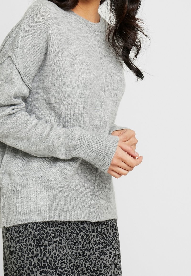 Femme portant un pull en maille gris doux à manches longues et une jupe imprimée léopard gris et noir, debout les mains jointes.