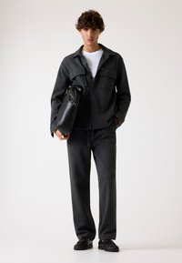 Giacca in misto lana grigia con tasche frontali, indossata su una camicia bianca. Pantaloni grigi abbinati e mocassini neri con dettagli testurizzati.