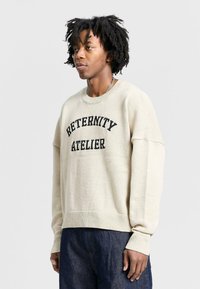 Beige knit sweater with black "RETERNITY ATELIER" text, wide cuffs, and ribbed hem. Worn over dark blue denim pants.