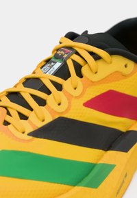 Baskets jaunes avec lacets jaunes, présentant des rayures rouges, noires et vertes, et une étiquette affichant un logo multicolore sur la languette.