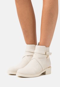Bottines beige clair en daim doux avec deux sangles croisées, une petite boucle sur le côté et un panneau arrière texturé, dotées d'un talon bloc.