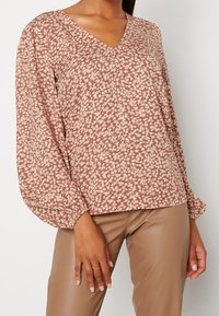 Blusa a maniche lunghe con scollatura a V e motivo floreale in beige e taupe. Tessuto morbido con taglio rilassato e polsini arricciati. Abbinata a pantaloni marroni.