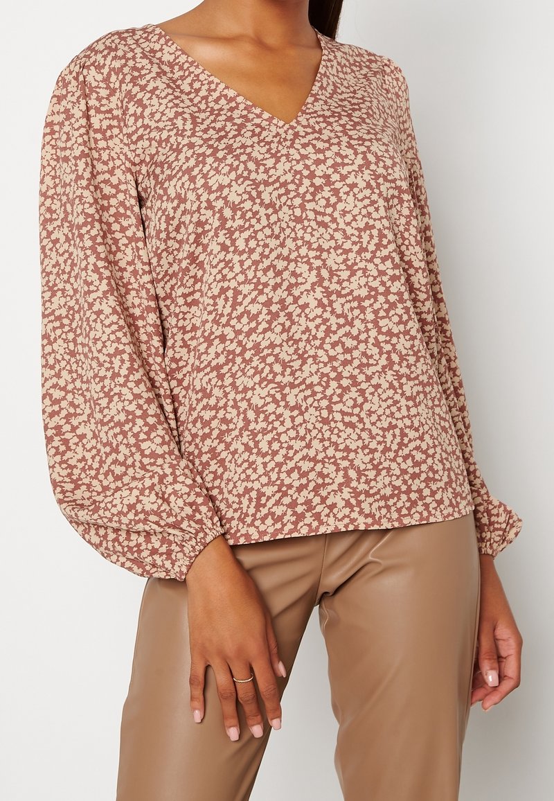 Blusa a maniche lunghe con scollatura a V e motivo floreale in beige e taupe. Tessuto morbido con taglio rilassato e polsini arricciati. Abbinata a pantaloni marroni.