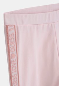 Pantaloni da jogging in cotone rosa chiaro con una fascia in coste in vita. Presentano una striscia decorativa laterale con la scritta "GUESS LOVE" e un logo a forma di cuore in rosa tenue.