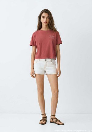 Ung kvinde står iført en rød t-shirt med korte ærmer, hvide denimbukser og brune studede sandaler mod en ensfarvet lys baggrund.