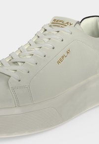 Basket basse blanche avec lacets blancs, marquage doré "REPLAY" sur le côté et la languette, et semelle épaisse texturée.