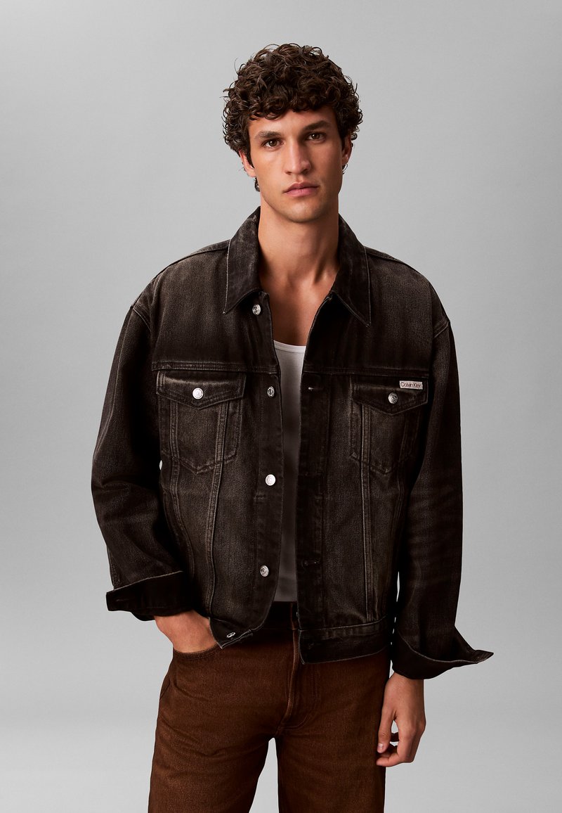 Schwarze Jeansjacke mit silbernen Metallbeschlägen, zwei Brusttaschen und einem lockeren Schnitt. Getragen über einem weißen Tanktop, kombiniert mit braunen Hosen.