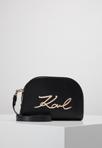 Borsa a tracolla in pelle nera con forma arrotondata, logo dorato "Karl", tracolla rimovibile, texture liscia e chiusura con zip nella parte superiore.