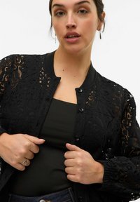 Veste bomber en dentelle noire avec un col rond, une fermeture à boutons et de longues manches froncées sur un haut sombre. Motif floral remarquable sur l'ensemble.