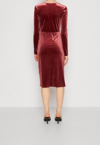 Femme portant une robe en velours à manches longues et longueur genou de couleur rouge profond, debout pieds nus sur un sol gris clair contre un mur uni.