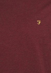 T-shirt in cotone bordeaux con una texture liscia, caratterizzato da un piccolo logo "F" giallo ricamato sul petto. Nessun motivo o dettagli aggiuntivi.