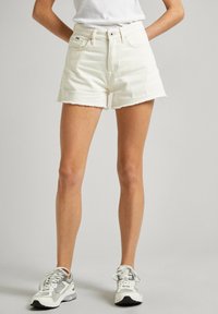 Shorts de mezclilla blanca con un dobladillo sin terminar, diseño de cinco bolsillos, cintura alta y cierre de botón. Combinados con zapatillas gris y blancas.