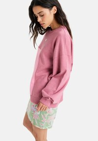 Lyserød bomulds-sweatshirt med sænkede skuldre og ribbede manchetter, parret med en pastelfarvet grøn nederdel med blomstermønster.