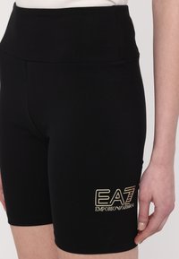 Črne raztegljive hlače s visokim pasom, s zlatim logotipom "EA7 Emporio Armani" na levem stegnu. Gladka tekstura, design do kolen.