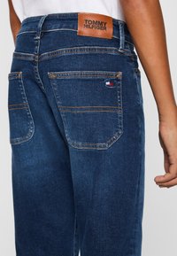 Jeans de mezclilla azul oscuro con costuras visibles, que presentan una etiqueta de cuero marrón con la inscripción "TOMMY HILFIGER" y un pequeño logotipo en el bolsillo trasero.