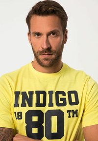 Camiseta de algodón amarilla con el texto negro en negrita "INDIGO 80 TM" exhibido de forma prominente en el pecho. Mangas cortas, corte estándar.