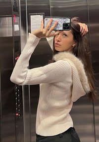 Vit ribbad tröja med en huva fodrad med päls. Fitted design, långa ärmar och mjuk textur. Reflekterad i en hisspegel.