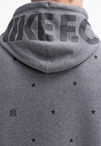 Šedá mikina s kapucí a černým nápisem "NIKE F.C." na kapuci. Na látce jsou hvězdné vzory v černé barvě. Měkký, strukturovaný materiál.
