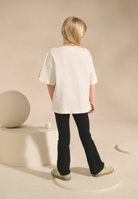 T-shirt blanc oversize, pantalons évasés noires côtelés, et baskets vertes avec des accents roses. L'arrière-plan présente des formes géométriques abstraites.