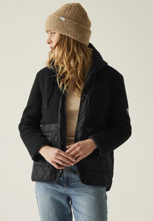 Mujer con gorro de punto beige, chaqueta acolchada negra con forro polar, camiseta beige de canalé y vaqueros azul claro, mirando hacia la izquierda con las manos entrelazadas.