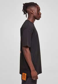 Sort bomulds T-shirt med korte ærmer, lige snit og et orange mærke i nederste venstre hjørne. Enkel og minimalistisk design.