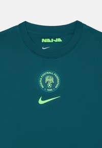Temno turkizna Nike majica z okroglo ovratnikom, teksturirana tkanina in zeleni grb Nogometne zveze Nigerije s orlom in nogometno žogo.