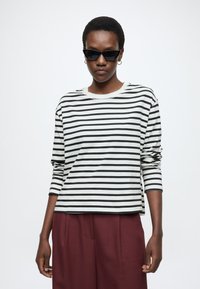 Csíkos, hosszú ujjú ing törtfehér és fekete színekben, kerek nyakkivágással és laza szabással, bordó nadrággal párosítva, modellt viselve.