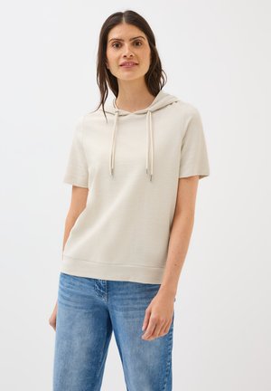 Haut à manches courtes beige clair en tissu texturé, avec un col à cordon et une coupe décontractée, associé à un jean en denim bleu.
