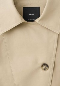 Material de palton bej cu textură netedă, guler mare și un singur nasture rotund lângă fante. Etichetă de marcă neagră în interior.