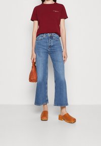 Mørkerød T-shirt med korte ærmer, højtsiddende lyseblå flagrende jeans, brune træskostøvler med nitter og en lille brun håndtaske. Glat tekstur.